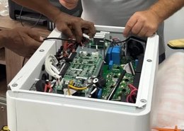 Mãos trabalhando nos conectores de um inversor aberto sobre a bancada.